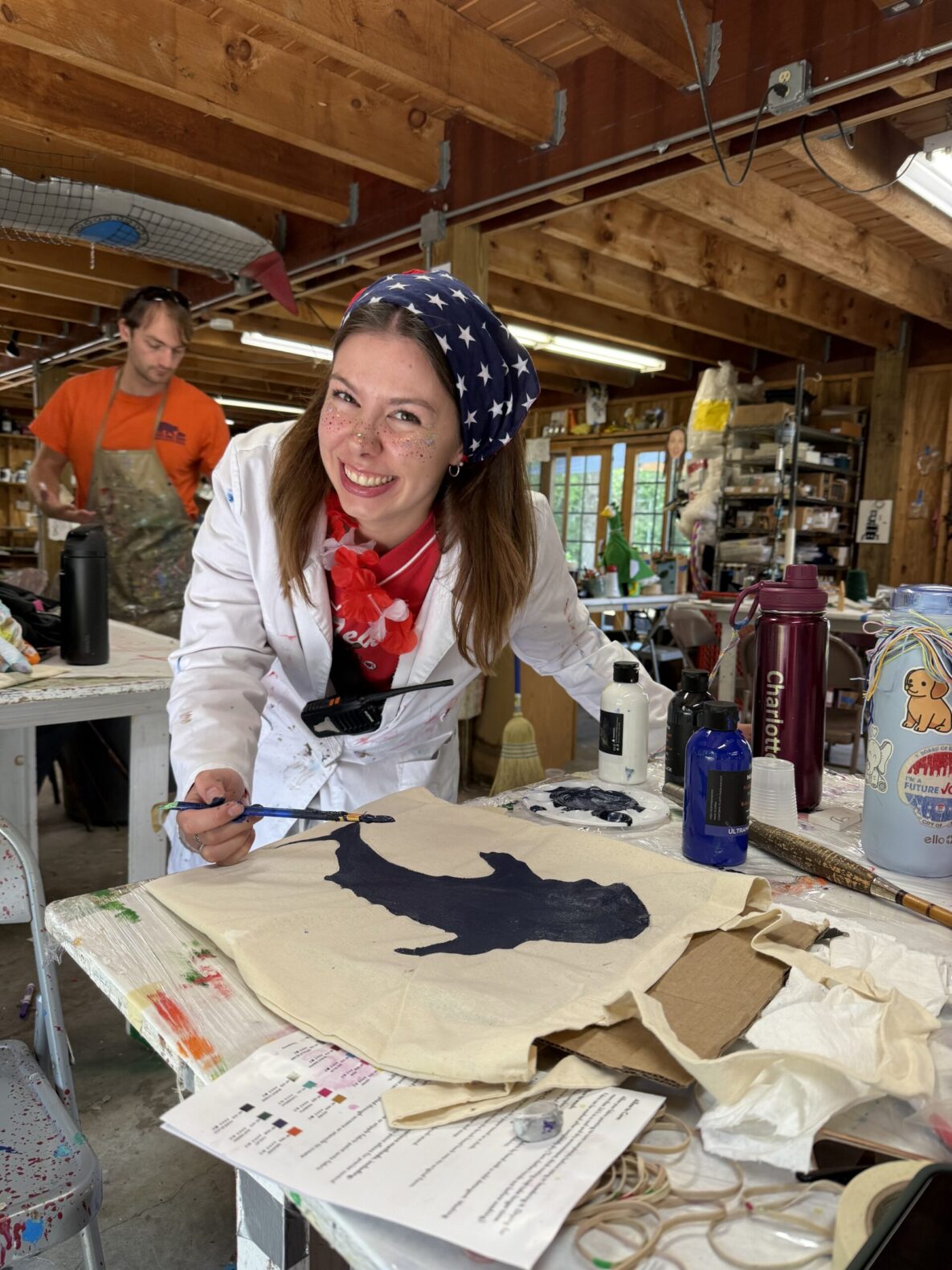Camp Connect USA - Woman in white shirt and red scarf at a craft table with various supplies painting.