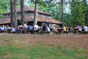 Camp Connect USA - Outdoor gathering with people seated on benches and chairs in a grassy area with trees and a building in the background.