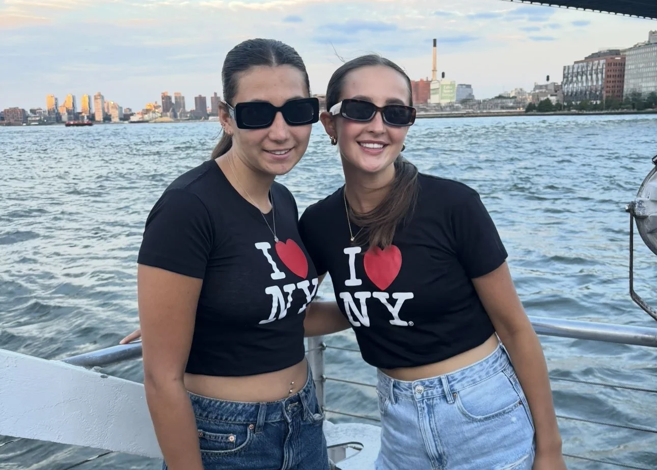 Camp Connect USA - Two people smiling on a boat, wearing sunglasses and matching black T-shirts with "I Love New York", with water and buildings in the background.